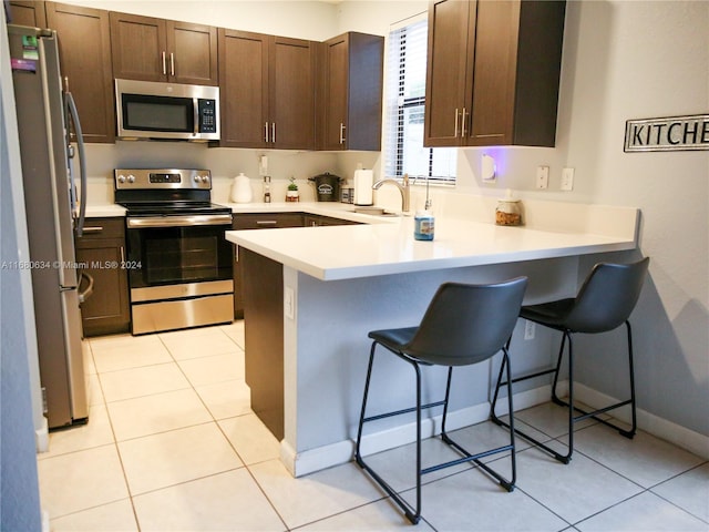 kitchen with kitchen peninsula, dark brown cabinets, light tile patterned floors, a kitchen bar, and stainless steel appliances