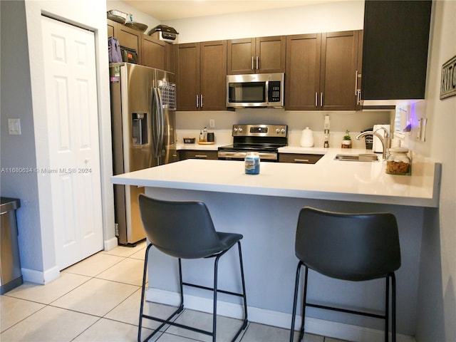 kitchen with a breakfast bar, dark brown cabinets, kitchen peninsula, stainless steel appliances, and light tile patterned floors