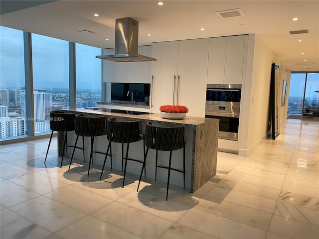 kitchen with a kitchen bar, island range hood, double oven, a large island, and expansive windows