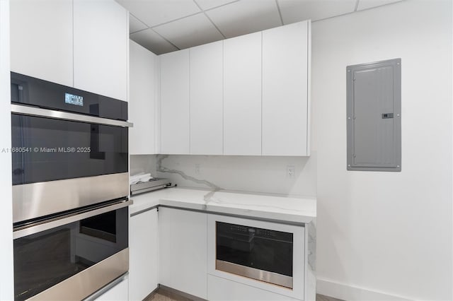 kitchen with decorative backsplash, light stone counters, double oven, electric panel, and white cabinetry