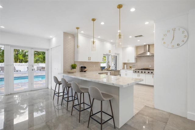 kitchen featuring a peninsula, wall chimney range hood, hanging light fixtures, and white cabinetry