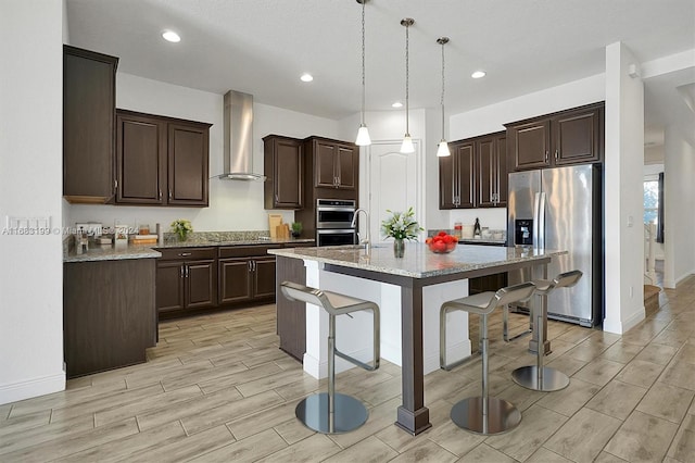 kitchen with wall chimney range hood, appliances with stainless steel finishes, dark brown cabinets, pendant lighting, and a center island with sink