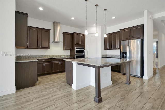 kitchen with wall chimney range hood, stainless steel appliances, pendant lighting, light hardwood / wood-style flooring, and a center island with sink