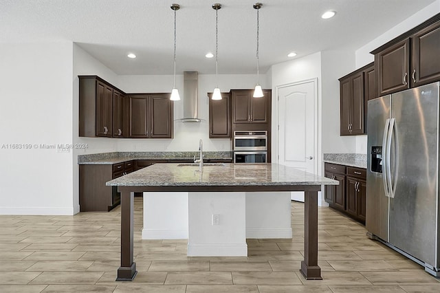 kitchen featuring wall chimney range hood, light stone counters, an island with sink, stainless steel appliances, and decorative light fixtures