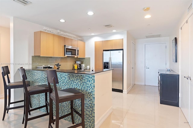 kitchen with light brown cabinets, kitchen peninsula, backsplash, light tile patterned flooring, and appliances with stainless steel finishes