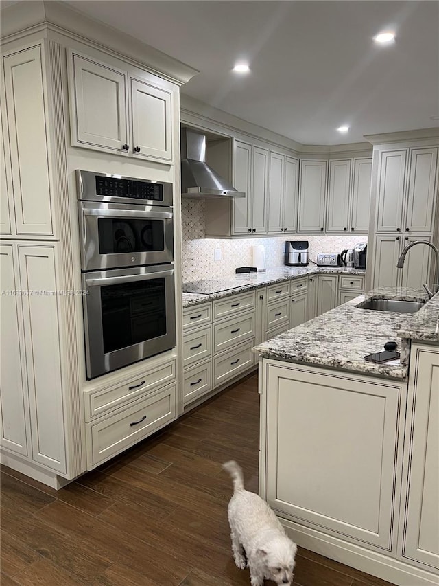 kitchen featuring light stone countertops, stainless steel double oven, dark wood-type flooring, and wall chimney range hood