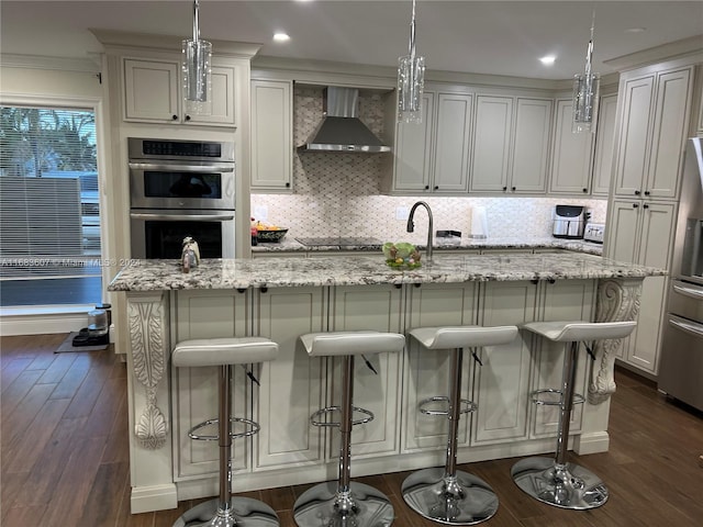 kitchen featuring wall chimney range hood, dark hardwood / wood-style flooring, an island with sink, decorative light fixtures, and a breakfast bar