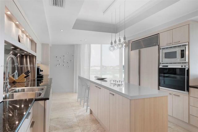 kitchen with light brown cabinets, a raised ceiling, a kitchen island, built in appliances, and decorative light fixtures