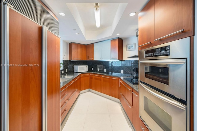 kitchen with tasteful backsplash, a tray ceiling, stainless steel double oven, paneled built in fridge, and dark stone counters