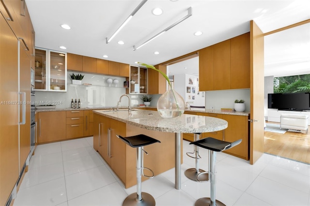 kitchen with light tile patterned floors, a kitchen breakfast bar, an island with sink, light stone countertops, and sink