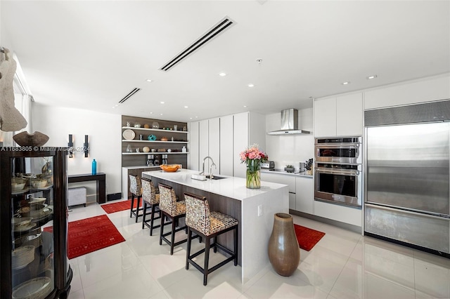 kitchen featuring wall chimney range hood, an island with sink, a kitchen breakfast bar, stainless steel appliances, and white cabinets