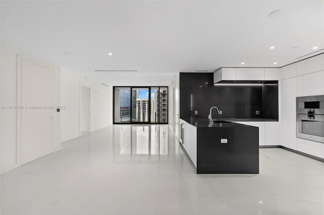 kitchen with sink, backsplash, stainless steel double oven, white cabinets, and a center island with sink