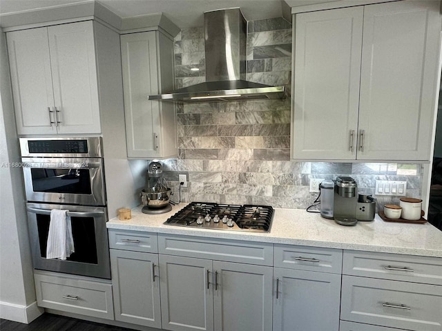 kitchen featuring light stone countertops, backsplash, stainless steel appliances, wall chimney range hood, and white cabinets