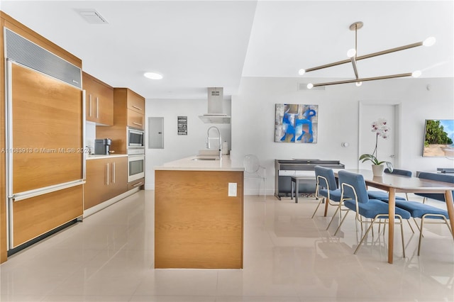 kitchen with wall chimney range hood, built in appliances, sink, decorative light fixtures, and light tile patterned floors