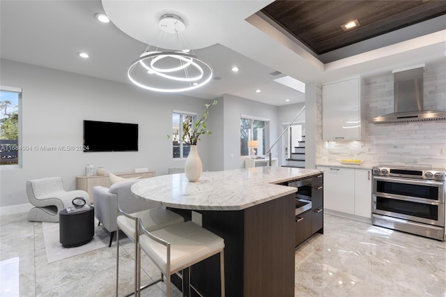 kitchen featuring white cabinets, tasteful backsplash, hanging light fixtures, range with two ovens, and wall chimney exhaust hood