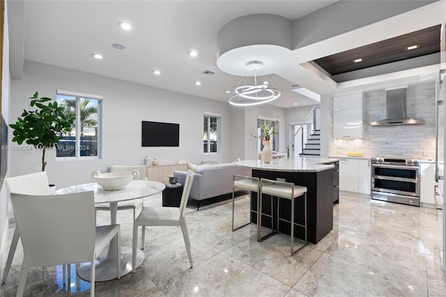 kitchen with wall chimney range hood, an island with sink, pendant lighting, white cabinetry, and electric stove