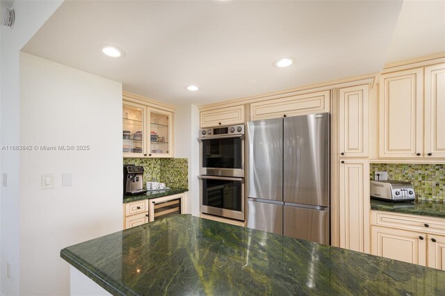 kitchen featuring dark stone counters, cream cabinetry, stainless steel appliances, beverage cooler, and backsplash