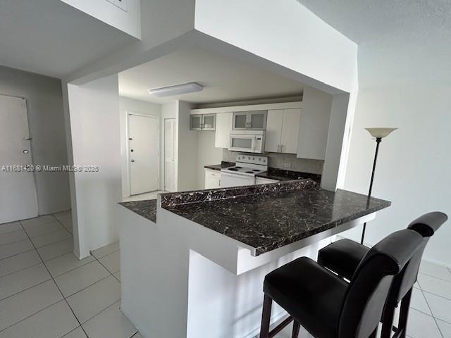 kitchen featuring light tile patterned floors, white cabinetry, kitchen peninsula, white appliances, and a breakfast bar