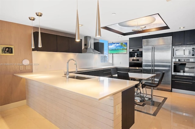 kitchen featuring wall chimney range hood, kitchen peninsula, a breakfast bar, built in appliances, and sink