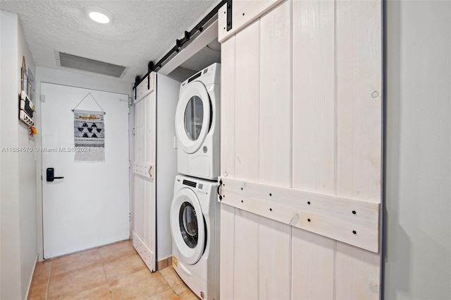 clothes washing area featuring stacked washer / drying machine, a textured ceiling, light tile patterned floors, and a barn door