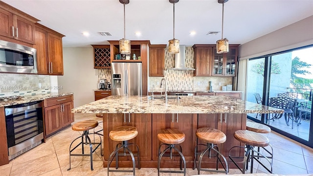 kitchen featuring hanging light fixtures, wine cooler, wall chimney exhaust hood, an island with sink, and appliances with stainless steel finishes