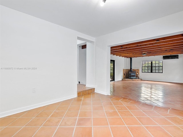 empty room with a wood stove, a wall mounted AC, beamed ceiling, and light tile patterned floors