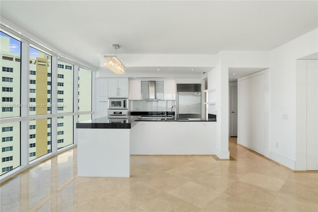 kitchen featuring built in appliances, pendant lighting, decorative backsplash, white cabinets, and wall chimney exhaust hood