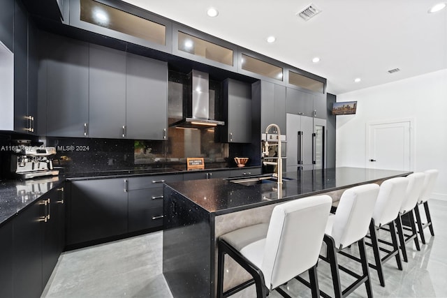 kitchen with decorative backsplash, dark stone counters, a breakfast bar, wall chimney range hood, and an island with sink