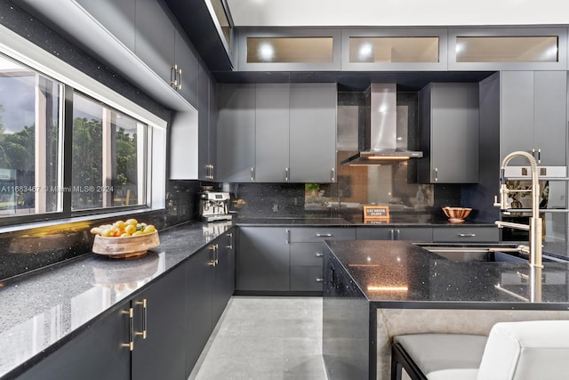 kitchen with gray cabinetry, sink, wall chimney range hood, backsplash, and dark stone countertops