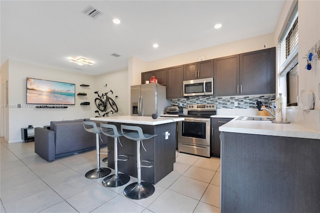 kitchen with a kitchen bar, appliances with stainless steel finishes, dark brown cabinets, light tile patterned floors, and a kitchen island