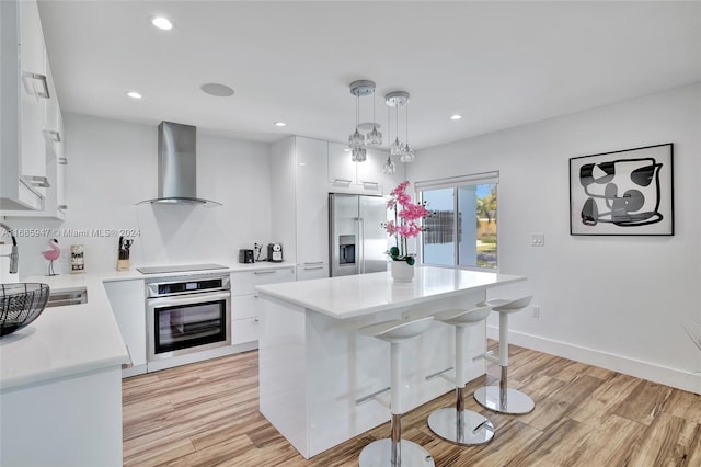 kitchen with wall chimney exhaust hood, hanging light fixtures, stainless steel appliances, light wood-type flooring, and white cabinets