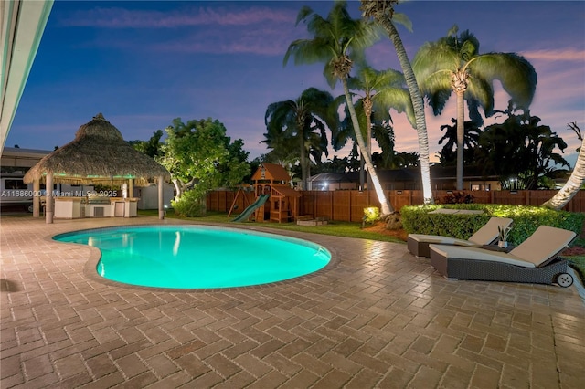 pool at dusk featuring a playground, a patio, an outdoor kitchen, and a gazebo