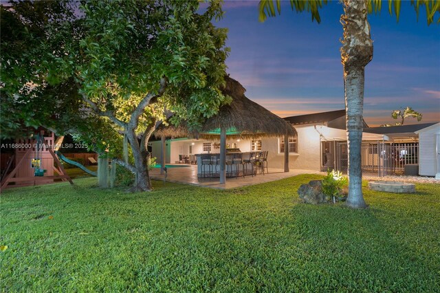 pool at dusk featuring an outdoor kitchen, a gazebo, a playground, and a patio