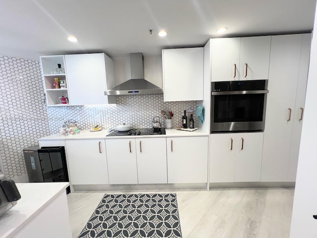 kitchen with black electric stovetop, wall chimney range hood, oven, light wood-type flooring, and white cabinets