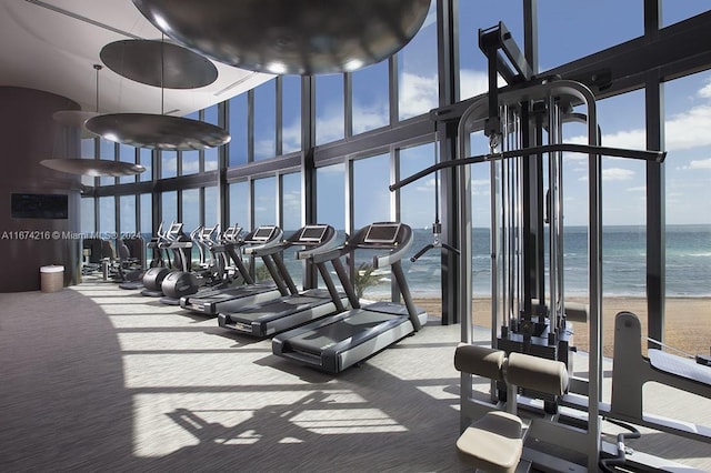 exercise room featuring carpet floors, a view of the beach, expansive windows, a water view, and a towering ceiling