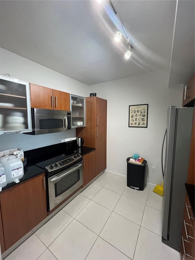 kitchen with brown cabinetry, dark countertops, appliances with stainless steel finishes, glass insert cabinets, and a textured ceiling