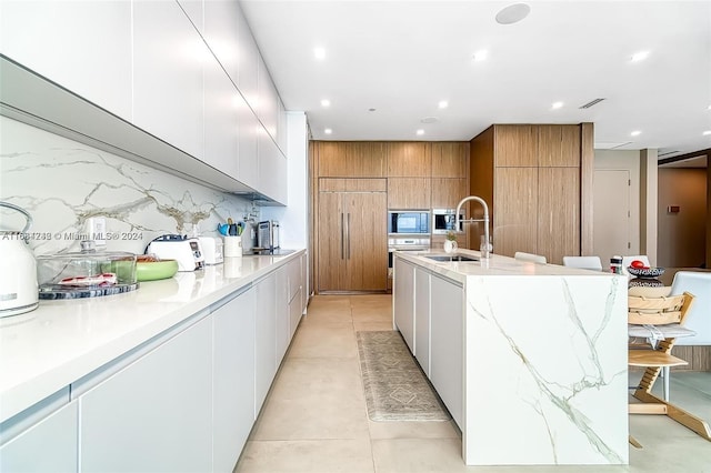 kitchen with tasteful backsplash, sink, white cabinetry, black microwave, and a kitchen island with sink