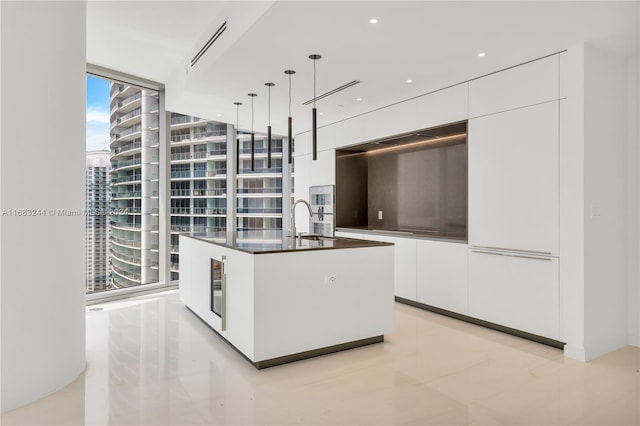 kitchen with a wall of windows, a kitchen island with sink, hanging light fixtures, and white cabinetry