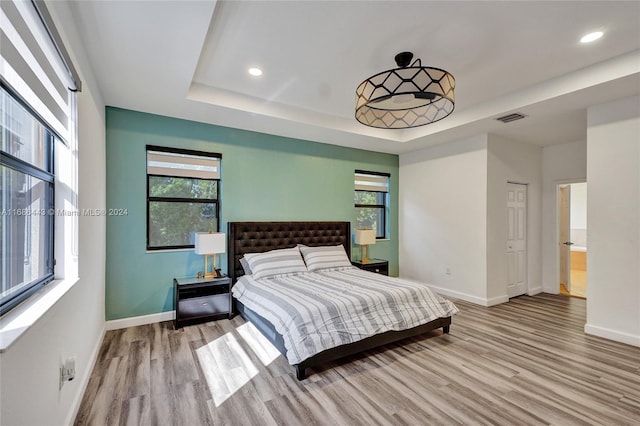 bedroom featuring light hardwood / wood-style floors, a raised ceiling, multiple windows, and ensuite bath