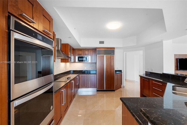 kitchen with tasteful backsplash, sink, built in appliances, a raised ceiling, and wall chimney exhaust hood