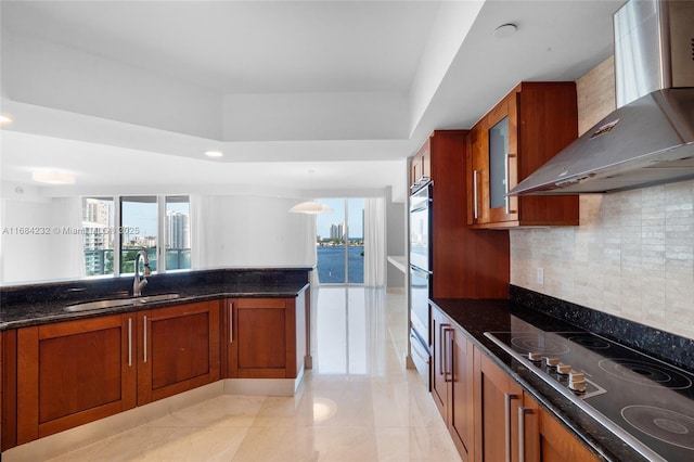 kitchen with wall chimney range hood, sink, gas stovetop, decorative light fixtures, and dark stone counters