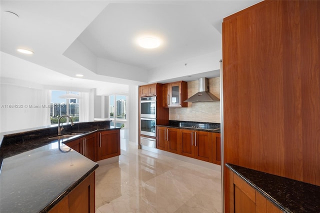 kitchen with dark stone countertops, double oven, black electric cooktop, decorative backsplash, and wall chimney exhaust hood