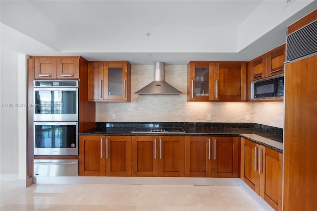 kitchen with tasteful backsplash, dark stone countertops, wall chimney range hood, and built in appliances