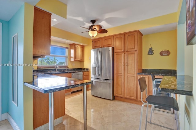 kitchen featuring appliances with stainless steel finishes, kitchen peninsula, ceiling fan, dark stone countertops, and a breakfast bar