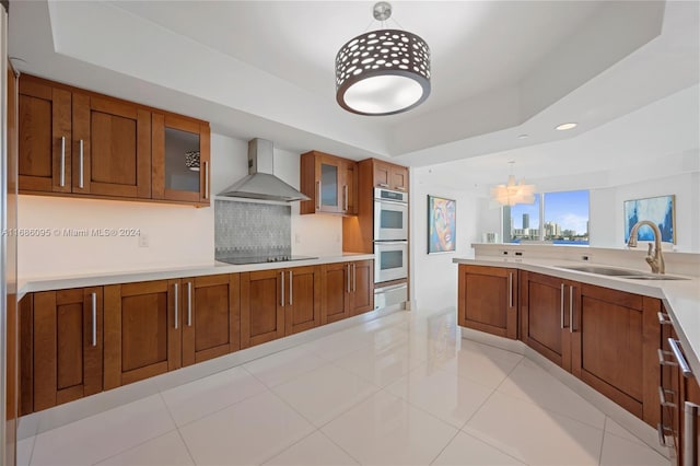 kitchen featuring sink, wall chimney exhaust hood, decorative light fixtures, stainless steel double oven, and a raised ceiling