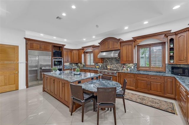 kitchen with a wealth of natural light, dark stone counters, stainless steel appliances, light tile patterned floors, and an island with sink