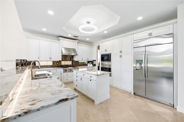 kitchen with light stone counters, range hood, white cabinetry, built in appliances, and sink