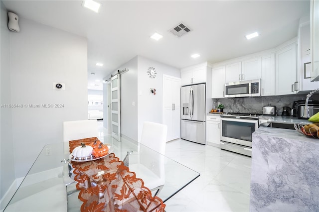 kitchen with white cabinets, backsplash, appliances with stainless steel finishes, a barn door, and light stone counters