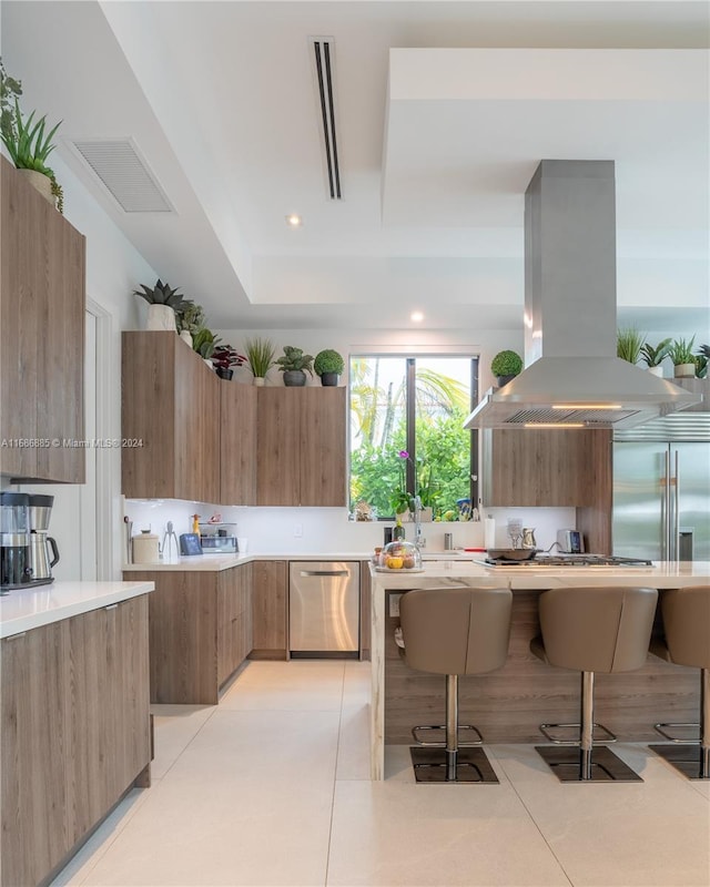 kitchen featuring light tile patterned flooring, a tray ceiling, island range hood, a kitchen bar, and stainless steel appliances