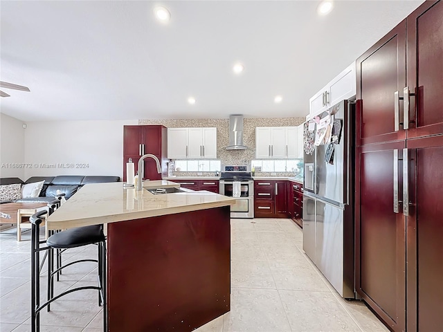 kitchen featuring appliances with stainless steel finishes, wall chimney exhaust hood, white cabinets, a breakfast bar area, and a kitchen island with sink